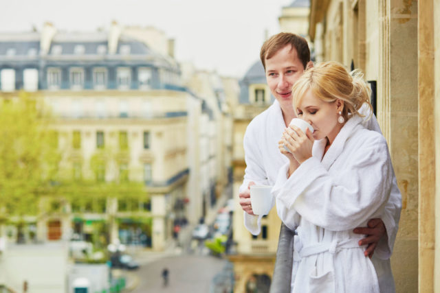  happy young couple in white bathrobes drinking coffee together on balcony with view to paris, france.